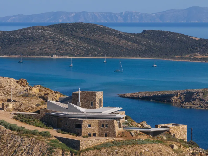 Hillside compound with stone walls and folded roof planes framing a bay with anchored sailboats