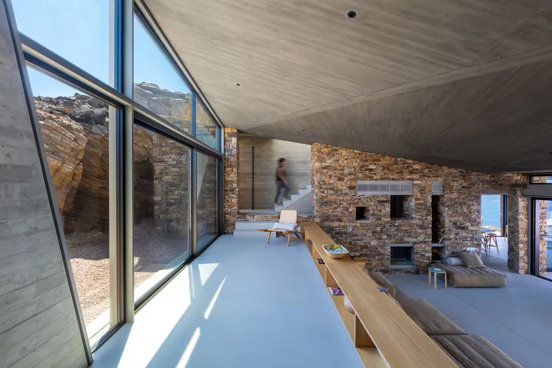 Interior living space with exposed stone walls, concrete ceiling, and glazed corner framing a figure in motion