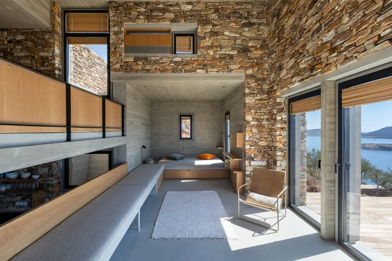 Double-height room with board-formed concrete walls, layered stone masonry, and timber shelving overlooking the bay
