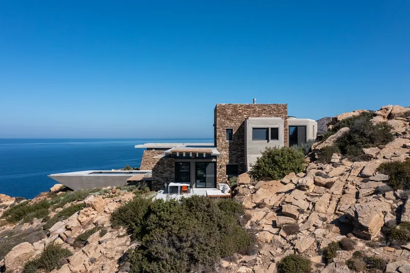 Stone and white plaster volumes perched on a rocky hillside overlooking the sea under clear skies