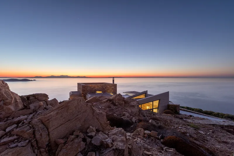 Stone volumes with illuminated windows perched on rocky outcrops at dusk above the water