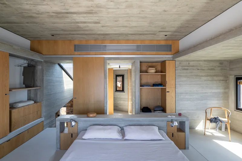 Bedroom with board-formed concrete walls, timber built-in wardrobes, and a doorway framing the passage beyond