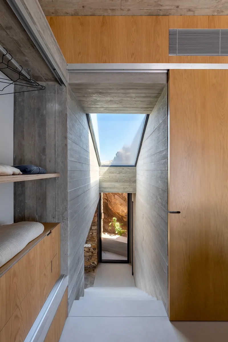 Corridor with angled skylight above board-formed concrete walls leading to a doorway with rock outcrop visible
