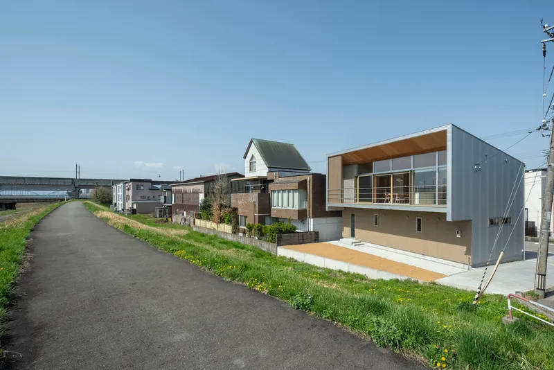 Diagonal view from adjacent path showing the house amid neighboring buildings and grassy embankment