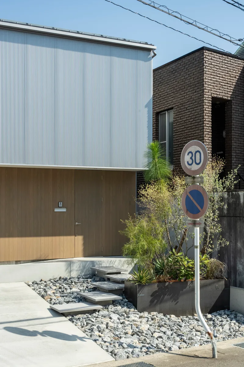 Street view showing corrugated metal and timber panel facades with planted beds along a gravel entrance path