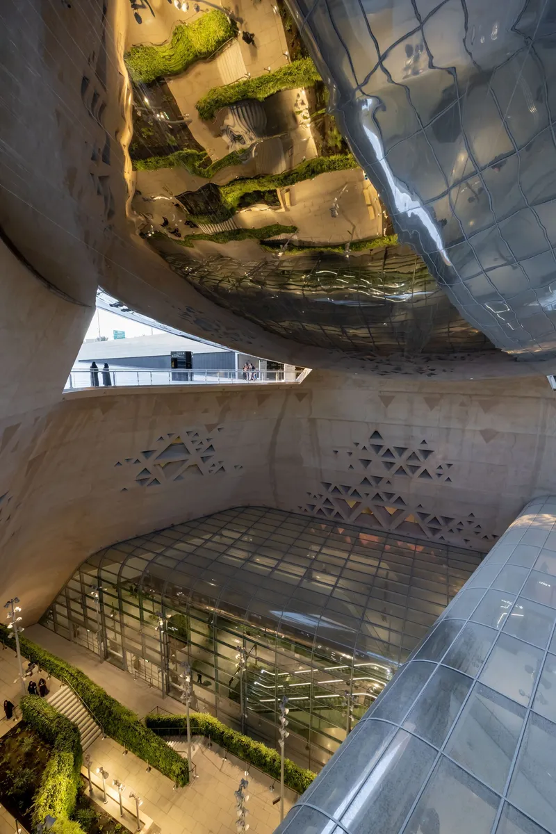 Upward view beneath the reflective metal canopy showing green planted terraces and curved glass elevator shaft with patterned stone base