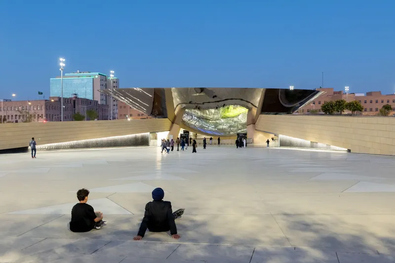 Plaza view at dusk with visitors gathered beneath the folded bronze canopy and illuminated entry pavilion