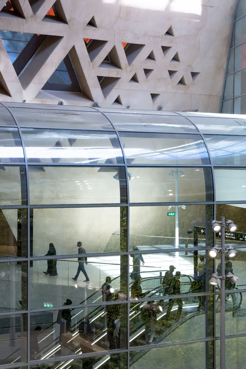 Interior view through curved glass escalator enclosure with concrete wall featuring triangular perforations and visitors descending below