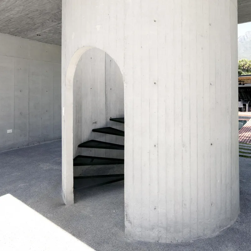 Cylindrical concrete core with arched opening enclosing a black spiral staircase in dappled sunlight