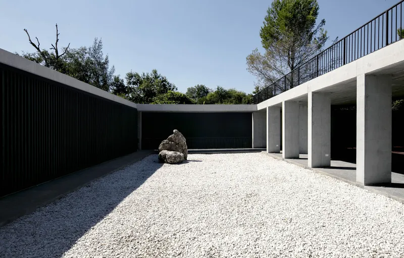 Gravel courtyard with a single boulder between concrete colonnade and black slatted wall