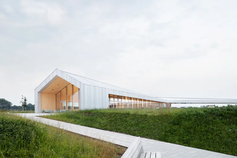 White corrugated metal facade with timber gable entrance beside a concrete walkway through grassy fields