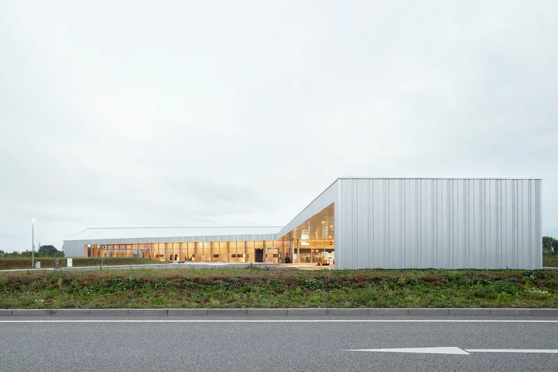 Long metal-clad volume with glazed timber pavilion viewed from the adjacent roadway at dusk