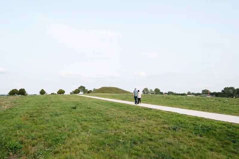 Concrete path crossing a green meadow with two people walking toward a distant mound