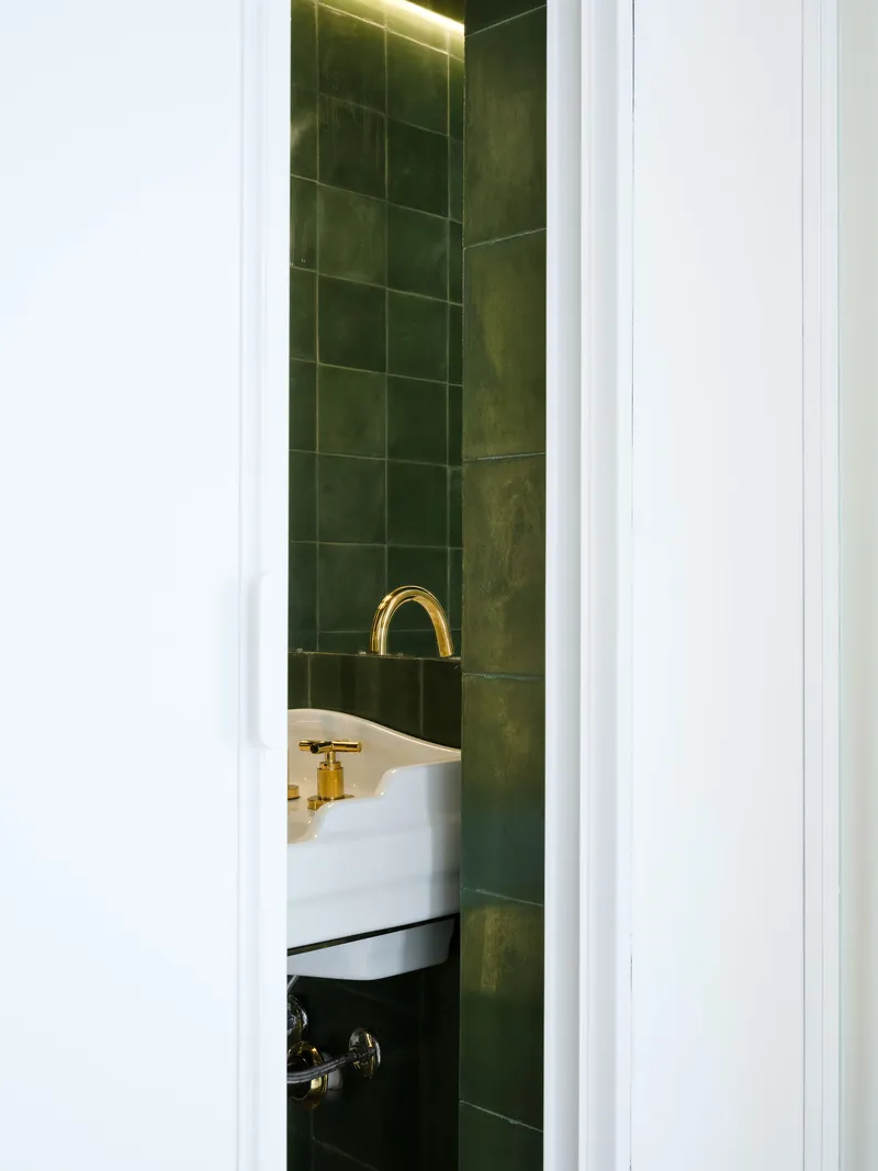 Glimpse through doorway into bathroom with dark green tile walls, brass faucet and concealed strip lighting