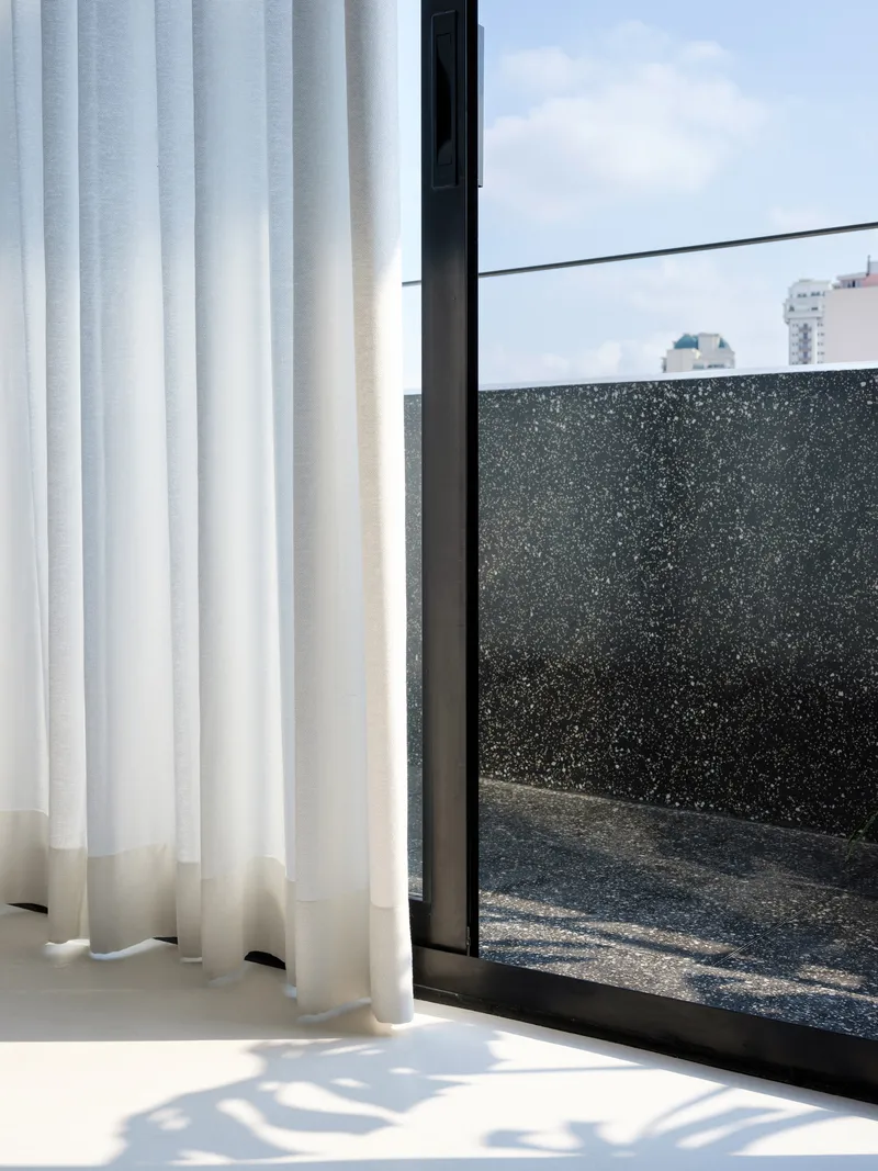 Floor-to-ceiling glazing with sheer curtain opening onto terrace with black terrazzo wall under daytime sky
