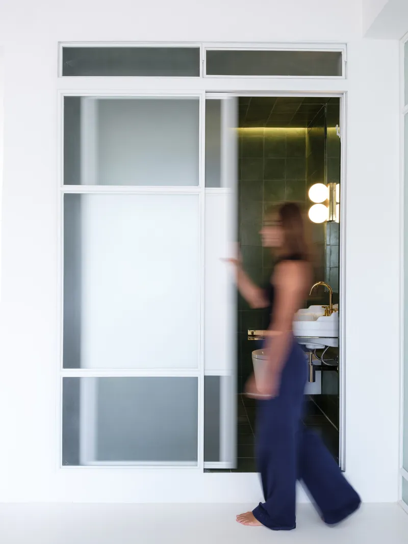 Figure in motion passing through the white-framed sliding door into the olive-tiled bathroom beyond