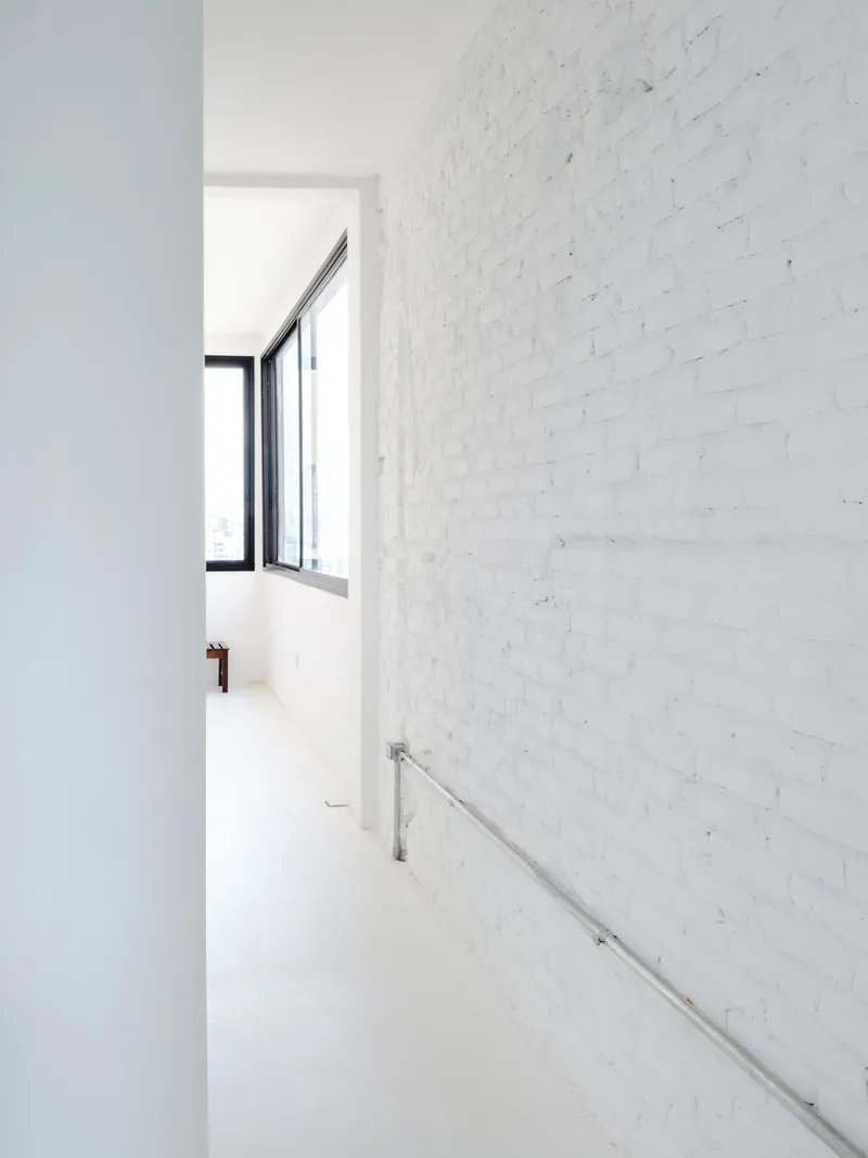 Narrow hallway with white-painted brick wall and stainless steel handrail leading toward a black-framed window
