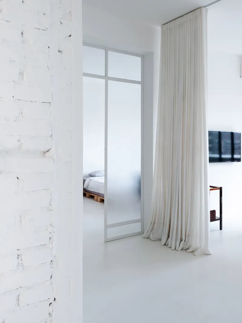 White interior with distressed brick wall, floor-to-ceiling sheer curtains and glimpse of bedroom beyond frosted glass partition