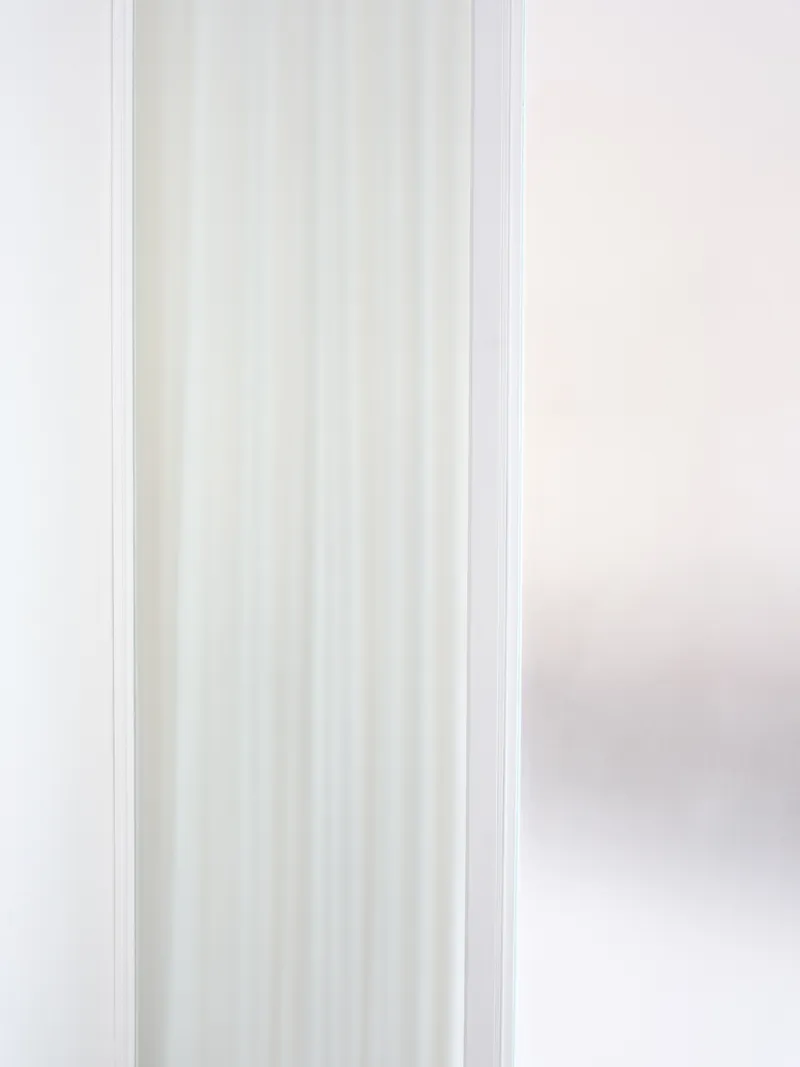 Close-up of translucent reeded glass panel framed in white metal alongside sheer curtain in soft daylight