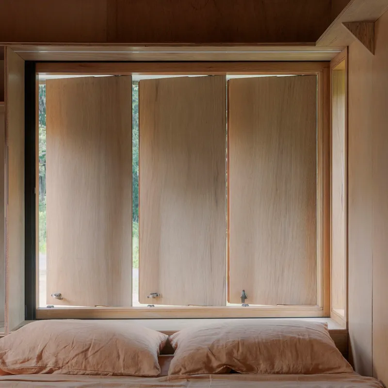 Bedroom window with three vertical timber panels framed in plywood above a linen-covered bed