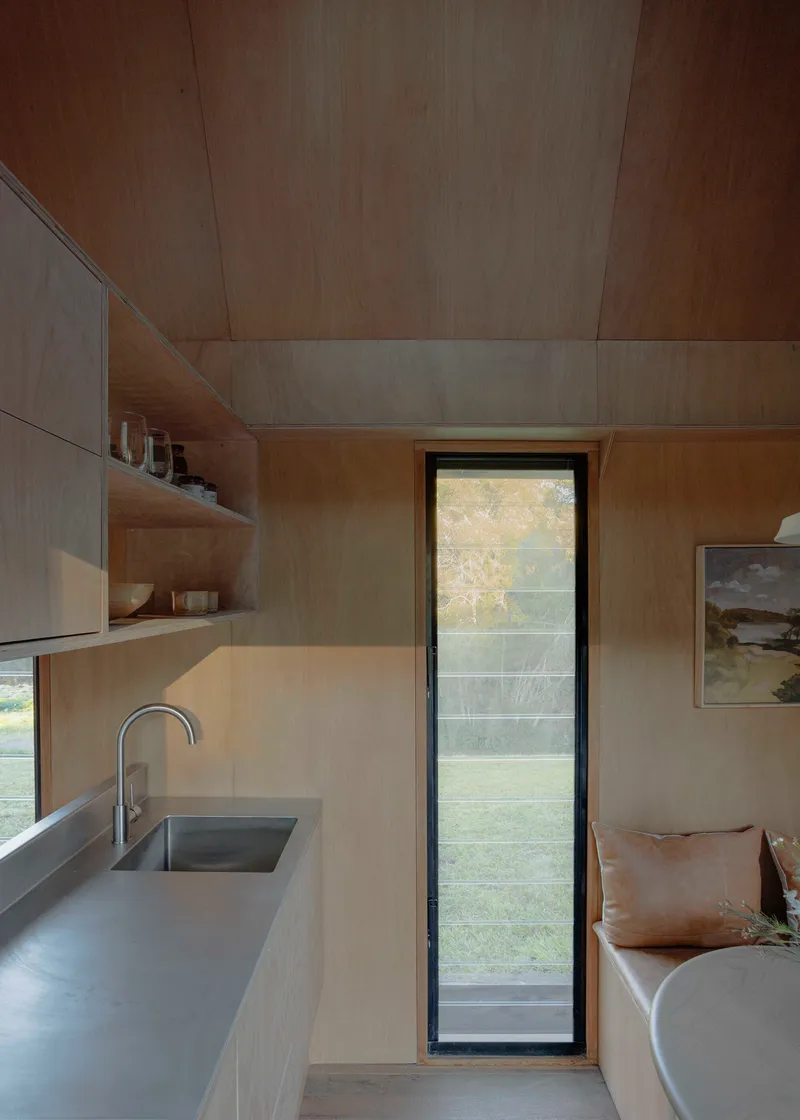 Plywood-lined kitchenette with integrated sink and narrow vertical window overlooking foliage outside