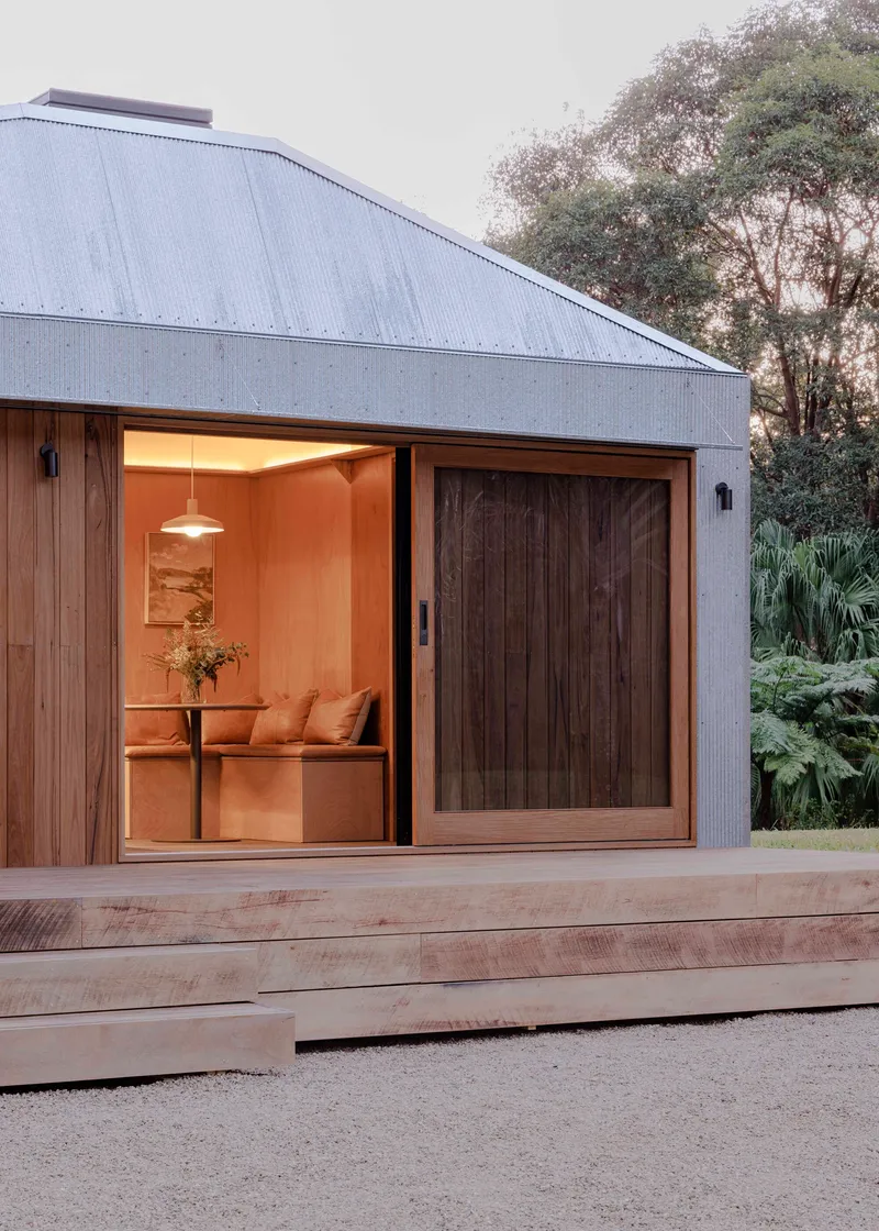 Corner of the pavilion at dusk showing sliding timber door and glowing interior under metal roof