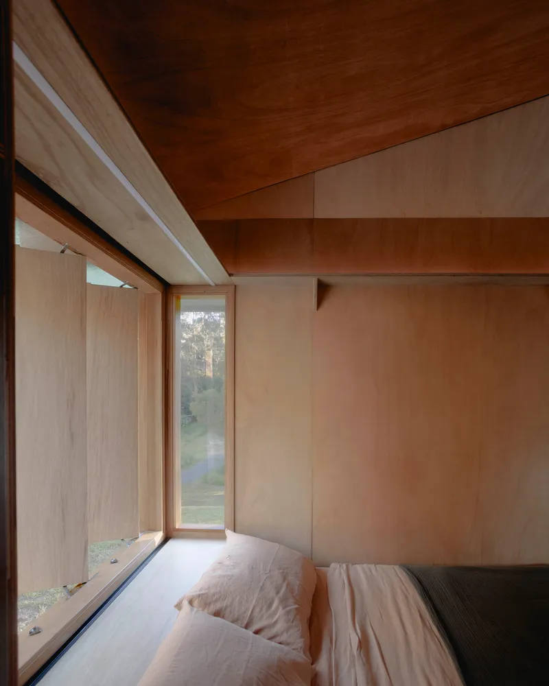Corner bedroom with plywood-lined walls and a low window seat overlooking the landscape
