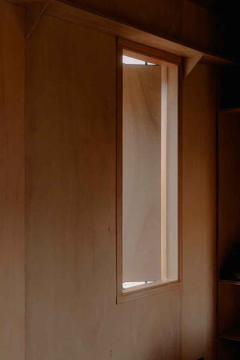 Tall narrow window set into plywood wall softly illuminated at dusk