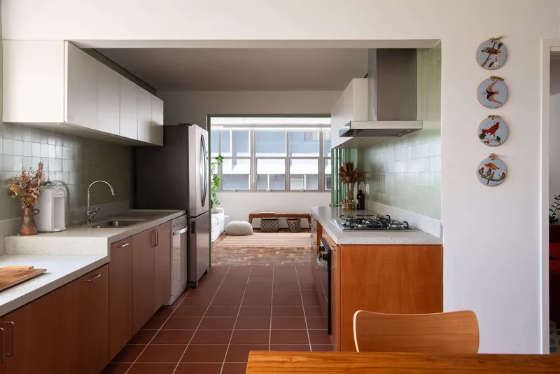 Galley kitchen with terracotta tile floor and green-framed window opening to an interior room