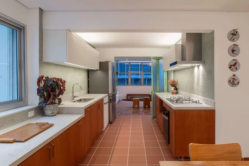 Galley kitchen with wood base cabinets, white upper cabinets, and terracotta tile floor leading to a blue louvered window