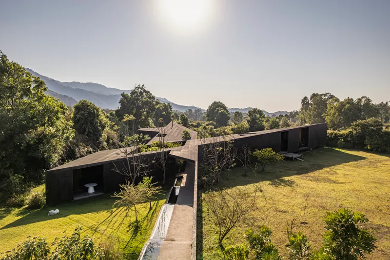 Aerial view of linear dark-clad volumes connected by a concrete bridge spanning a sloped lawn with mountains beyond