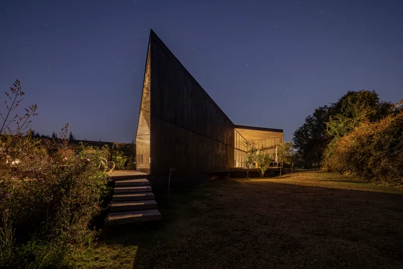 Elevated dark timber volume with illuminated glass pavilion beside it under a starlit night sky