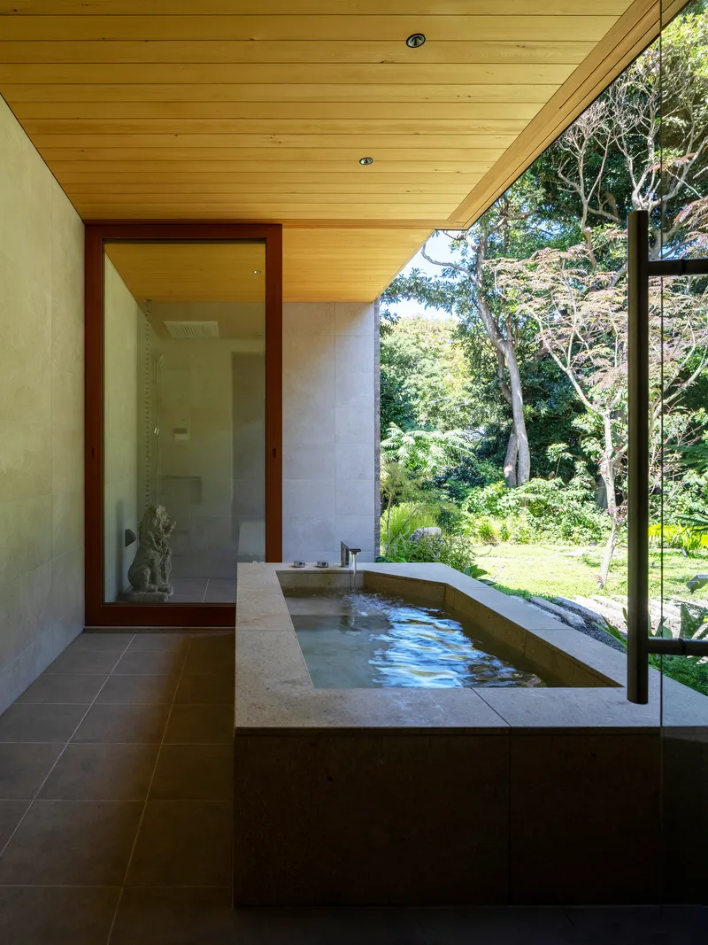 Bathroom with sunken concrete tub beside floor-to-ceiling glass overlooking garden and trees