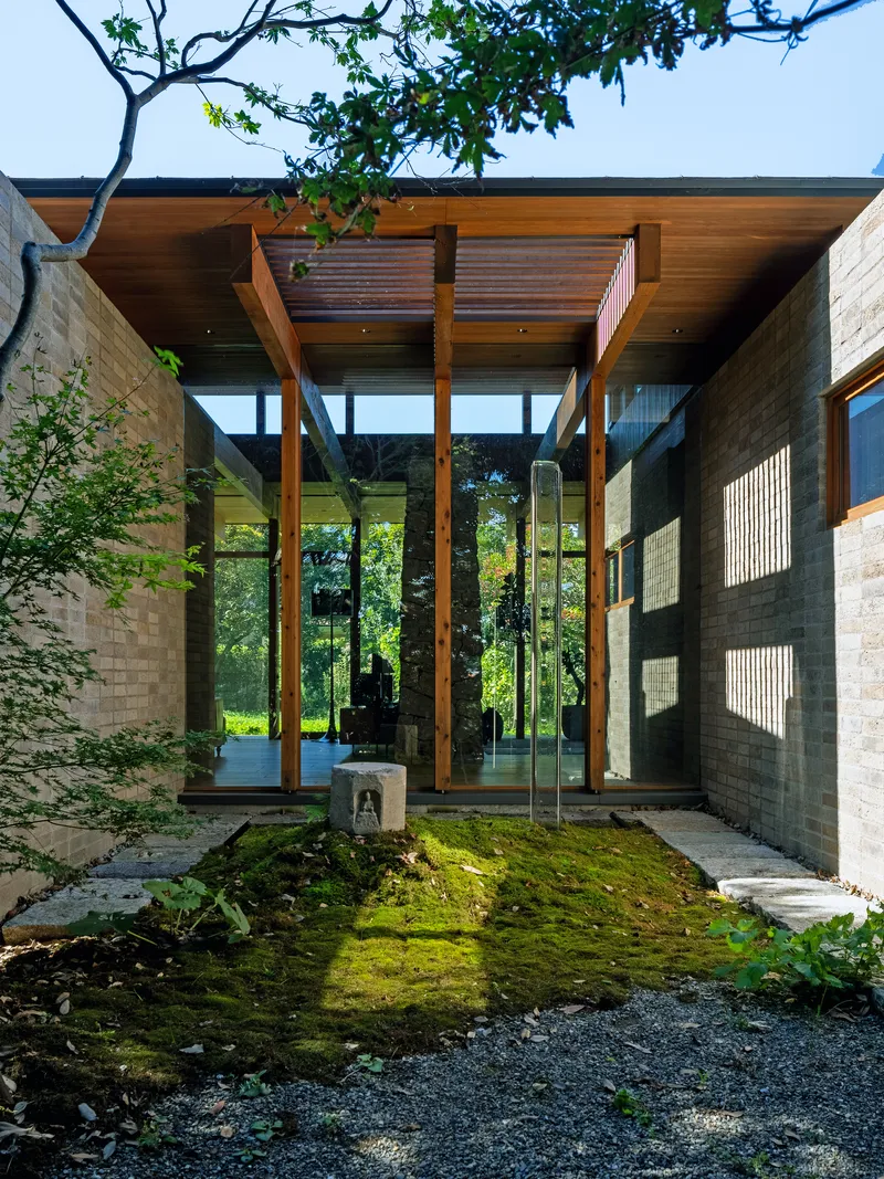 Glazed entrance pavilion with timber beams and slatted canopy framed by moss garden and stone pavers