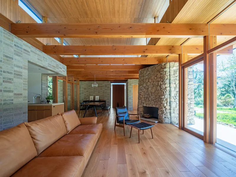 Living room with exposed timber beams, stone fireplace wall and full-height glazing opening to the lawn