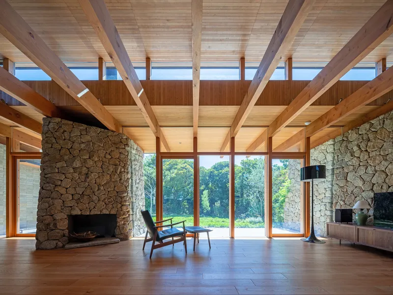 Stone fireplace wall and floor-to-ceiling windows framing views of trees beneath a timber beam ceiling