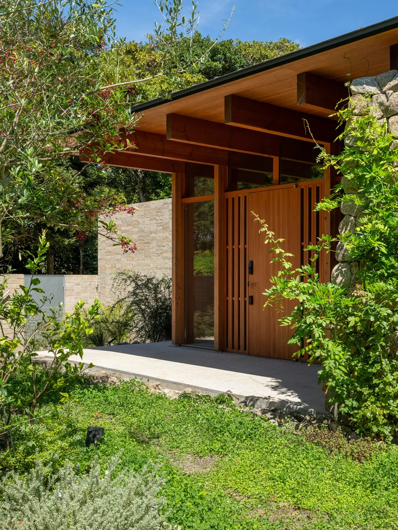 Timber entry pavilion with exposed beams and vertical slat door surrounded by dense vegetation