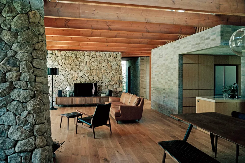 Living room with fieldstone fireplace wall and exposed timber ceiling at dusk