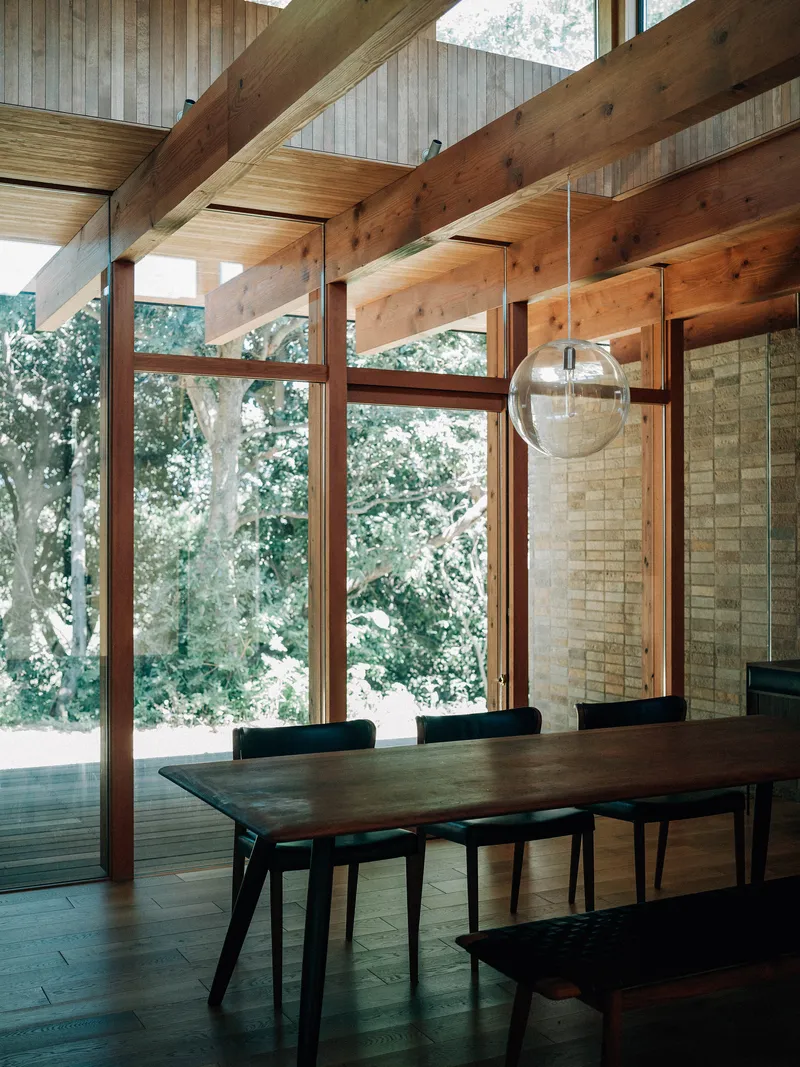 Dining table beneath timber ceiling and glass pendant with stone wall and forest view beyond