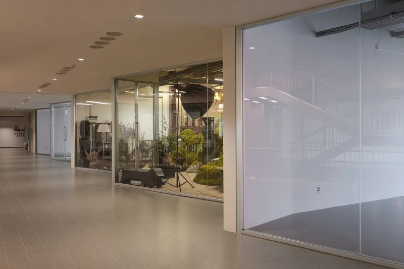 Glass-walled corridor with frosted and clear panels looking into a planted display area with studio lighting