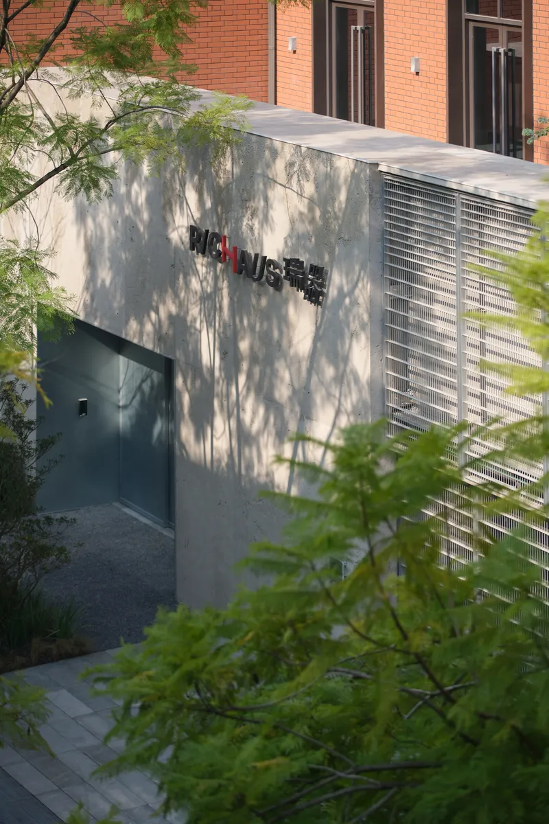 Concrete entry volume with perforated metal screen panel and recessed doorway framed by deciduous trees