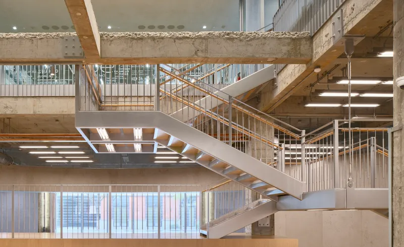Multi-level atrium with open timber and steel staircases between exposed concrete beams and white balustrades