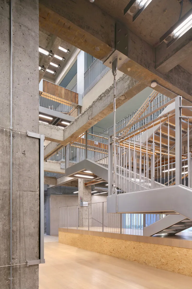 Multi-level interior atrium with exposed concrete columns, timber beams, and metal staircases under fluorescent lighting