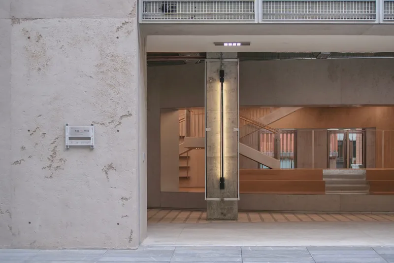 View through concrete portal showing timber volume with metal railing and exposed cylindrical column with wall-mounted light