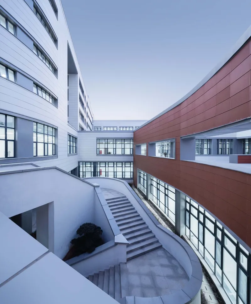 Curved staircase descending into a sunken courtyard between white and terracotta panel facades with glazed bridges