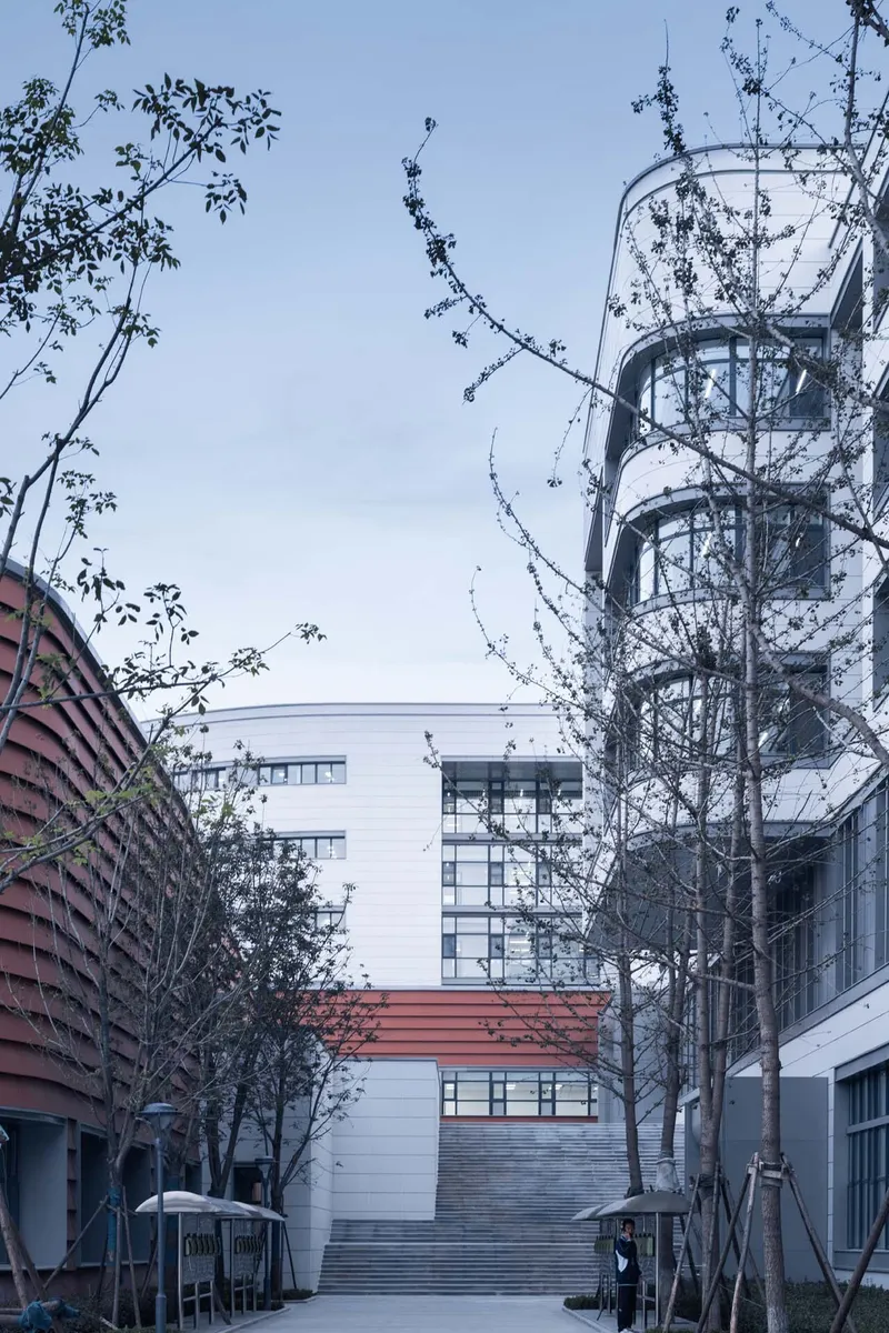 Pedestrian path through young trees leading to a cylindrical glass tower and terracotta louvered volume