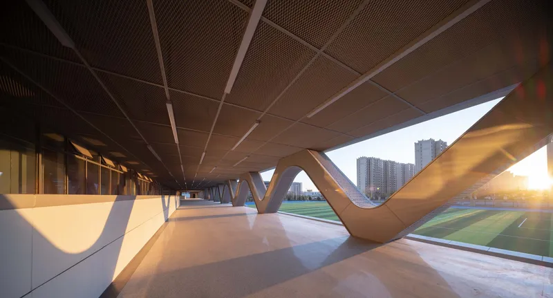 Covered colonnade with V-shaped columns and perforated wood ceiling panels at sunset