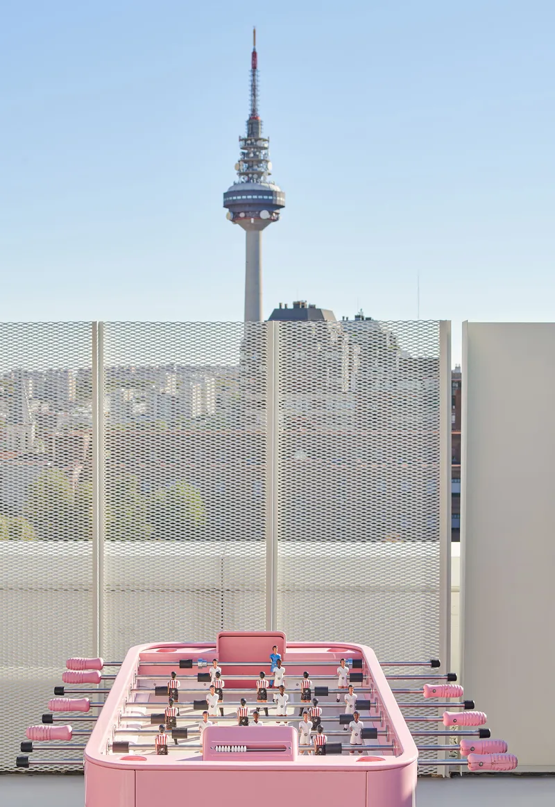 Pink foosball table on rooftop with perforated metal screens and telecommunications tower in distance