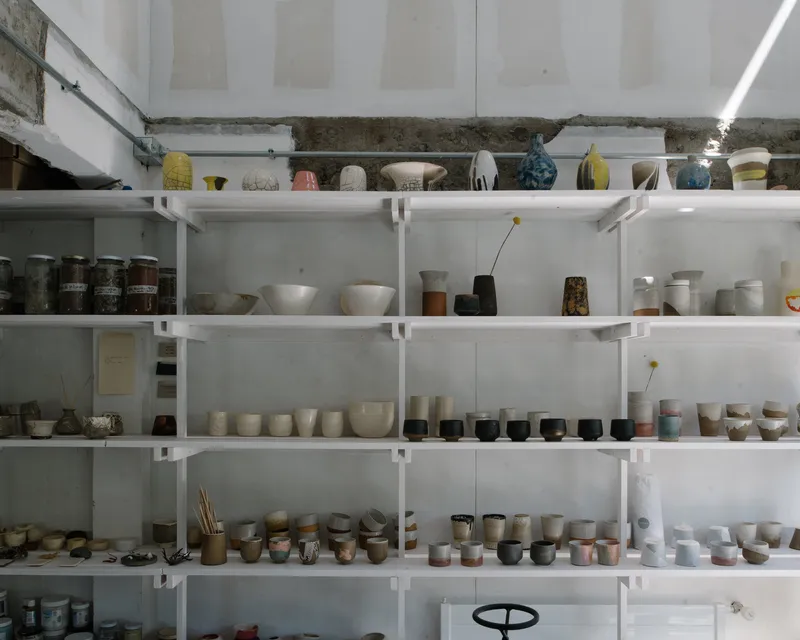 White metal shelving filled with ceramic vessels and pottery beneath a raw concrete beam