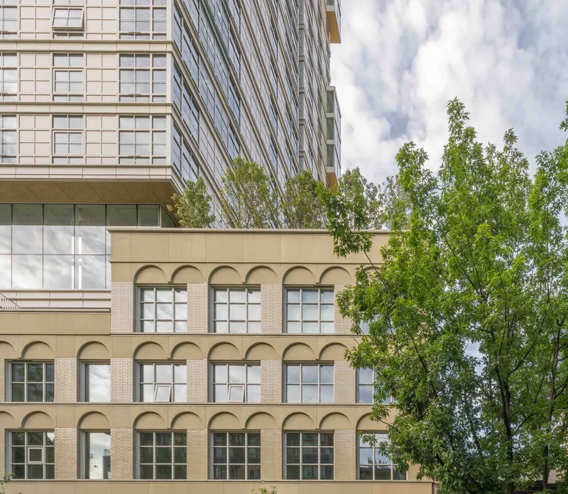 Arched window facade in beige brick below a cantilevered glass tower on a cloudy day